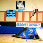 GO BIG BMX AIR Show assembly in school gym with GREGORY HUSKIES banner, BMX rider performing mid-air stunt over inflatable ramp