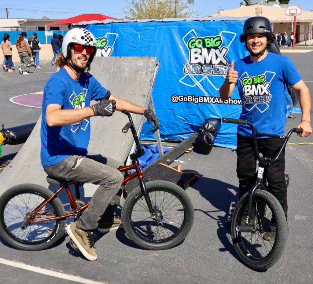 Two BMX riders posing with bikes in front of Go Big BMX Air Show banner promoting stunts and anti-bullying messages